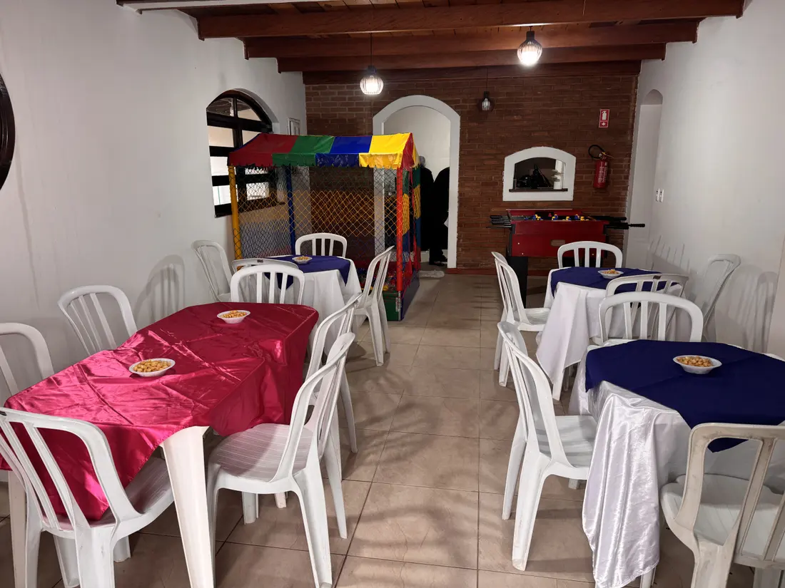 Espaço coberto com mesas de toalhas coloridas e, ao fundo, a piscina de bolinhas e o pebolim