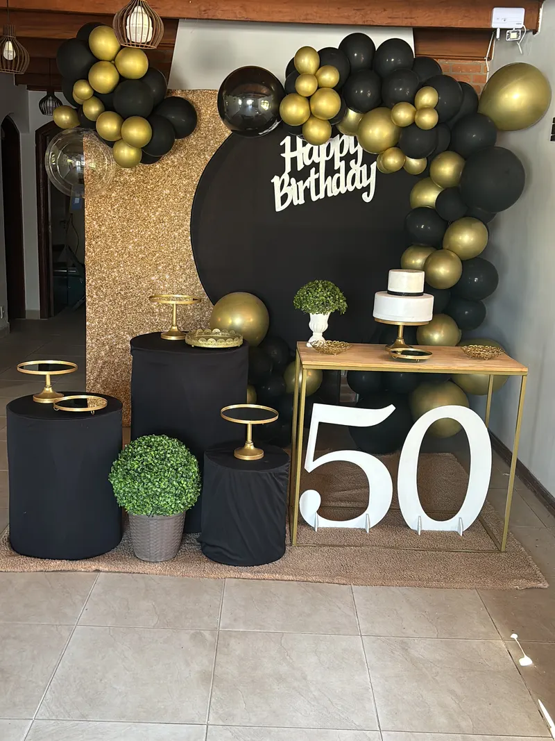 Mesa do bolo de 50 anos com painel preto “Happy Birthday”, balões pretos e dourados e o número 50