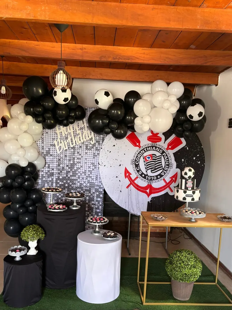 Festa com tema de futebol, balões pretos e brancos, bolas de futebol, painel com o escudo do time e bolo de futebol